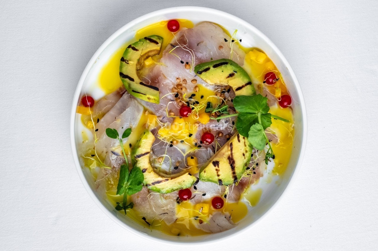 Assiette de carpaccio de poisson cru avec avocat grillé, groseilles rouges, pousses et sauce jaune.
