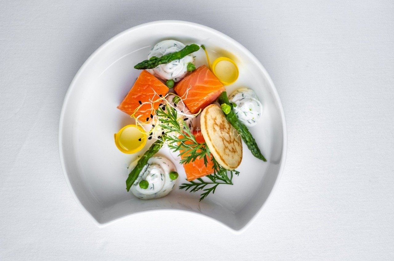 Assiette gastronomique avec saumon, asperges, crème aux herbes, crêpe et décorations végétales sur fond blanc.