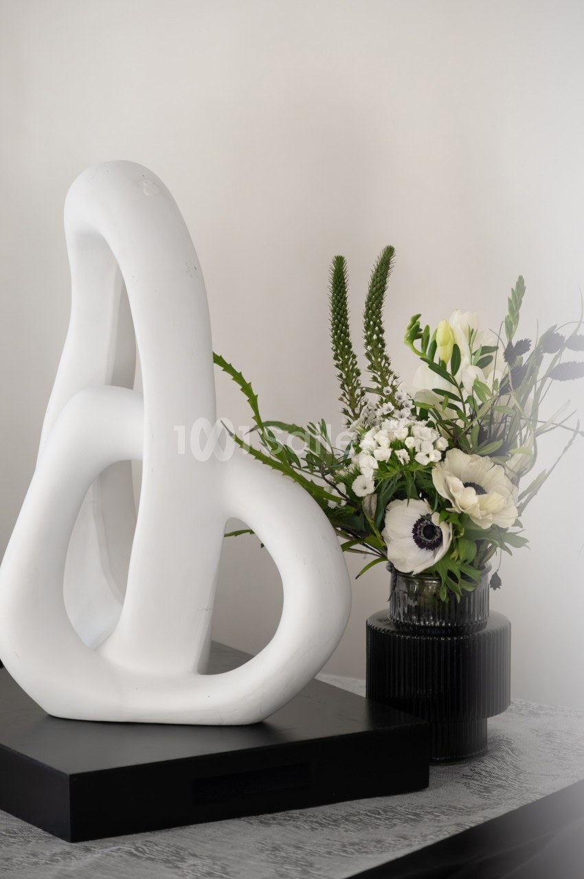 Sculpture blanche abstraite sur un socle noir, accompagnée d'un vase noir contenant un bouquet de fleurs blanches et vertes.