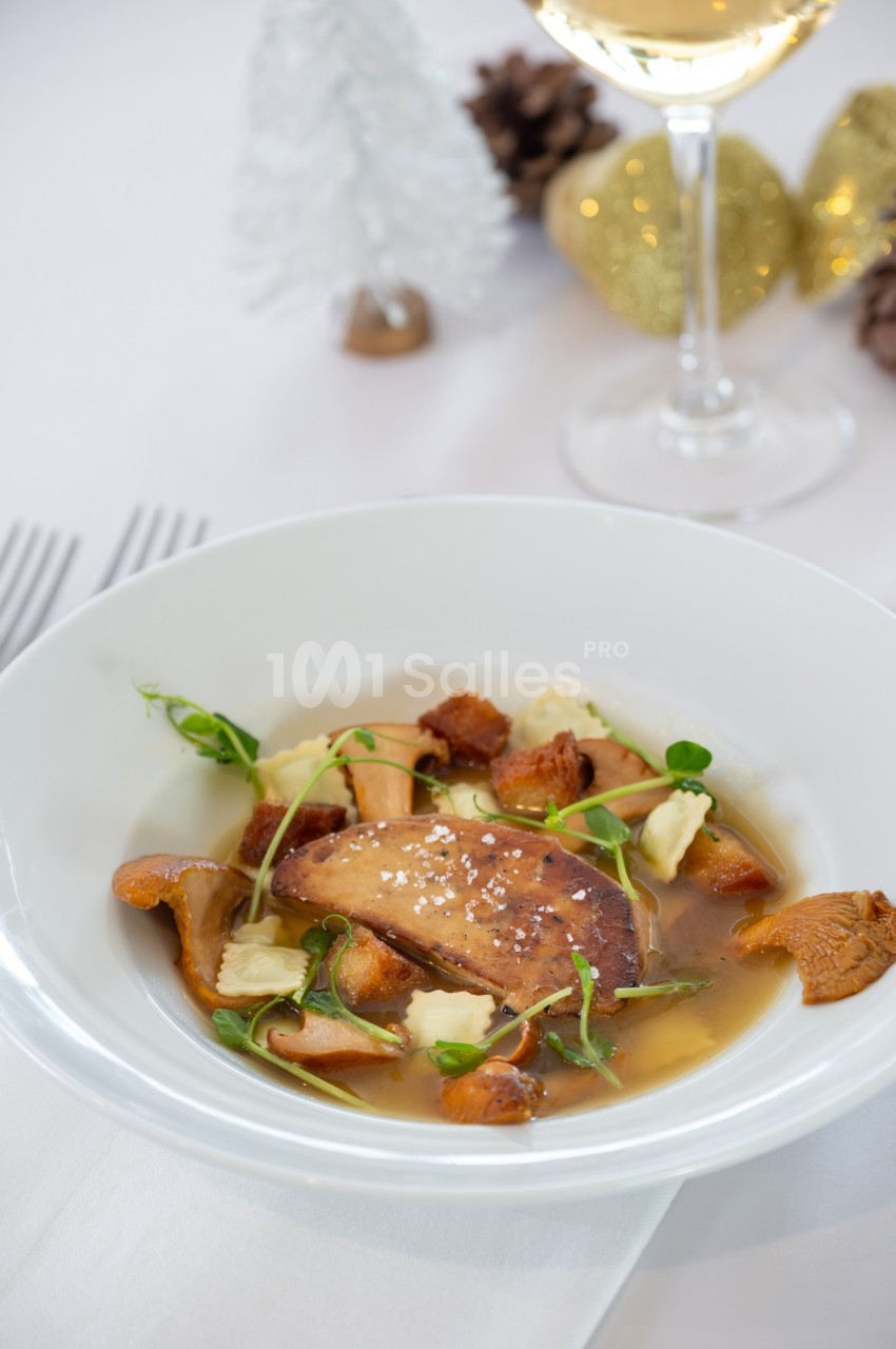 Assiette de foie gras poêlé avec champignons, herbes fraîches et bouillon, servie sur une table élégamment dressée.