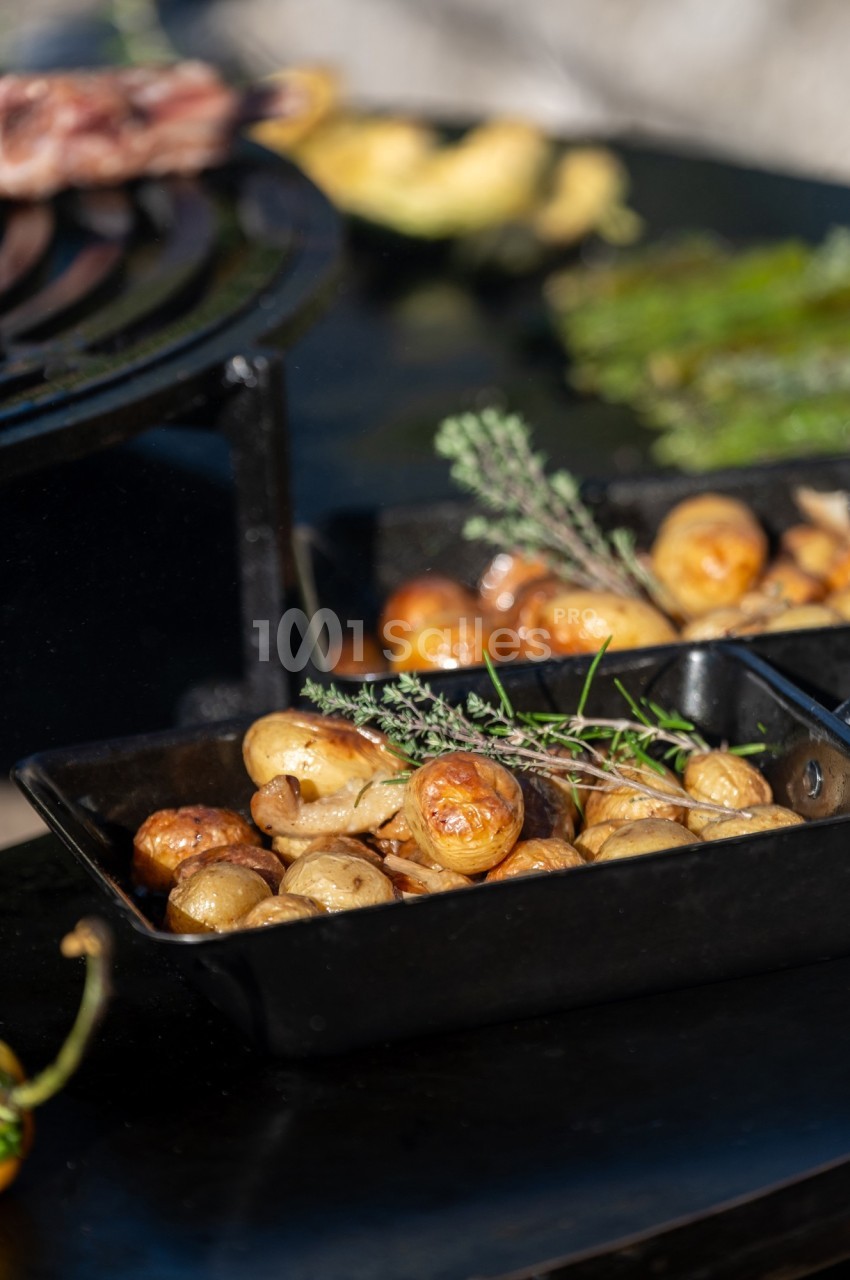 Pommes de terre rôties dans un plat noir, garnies de branches de thym, posées sur une table près d'un barbecue.