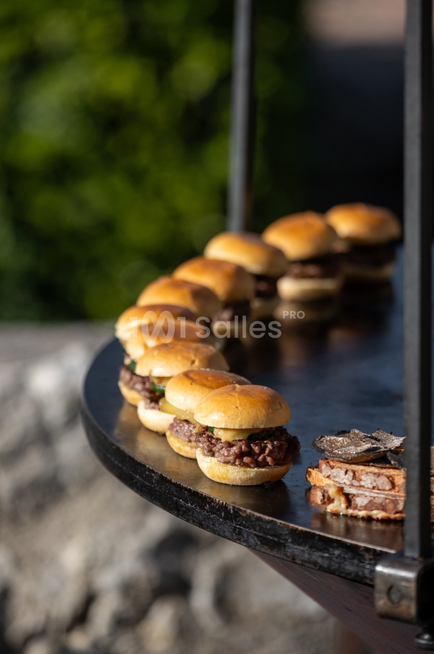 Mini-burgers alignés sur une table ronde en extérieur, avec des tranches de pain garnies de truffes au premier plan.