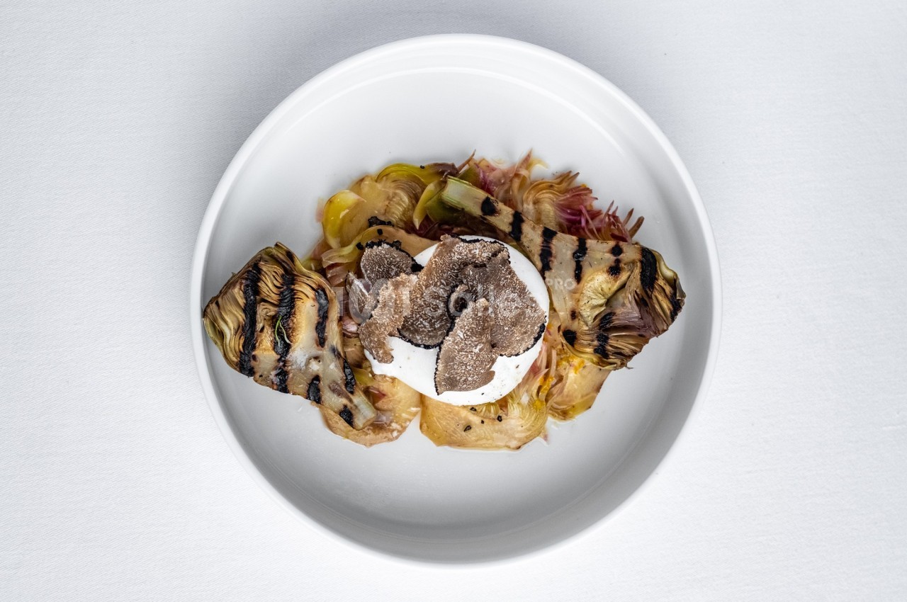 Assiette contenant des artichauts grillés, une burrata et des tranches de truffe noire, présentée sur une nappe blanche.