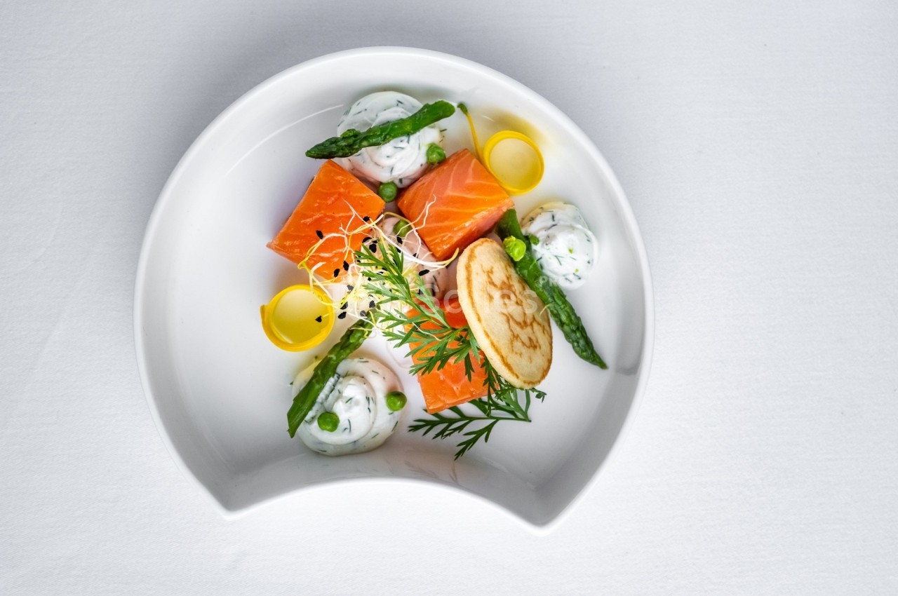 Assiette gastronomique avec saumon, asperges, crème aux herbes, crêpe et décorations végétales sur fond blanc.