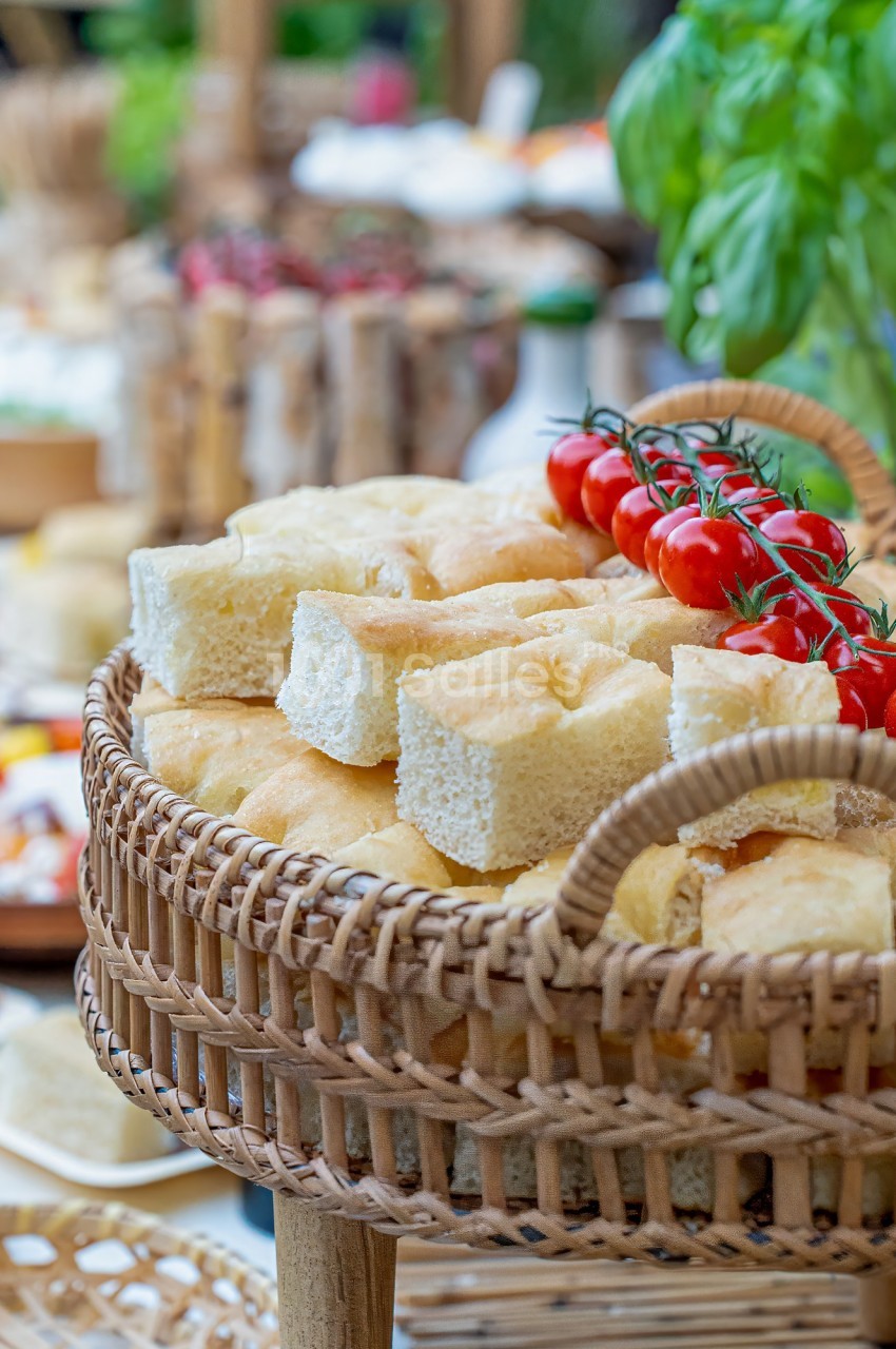 Panier en osier contenant des morceaux de focaccia et des tomates cerises sur une table garnie de mets variés.