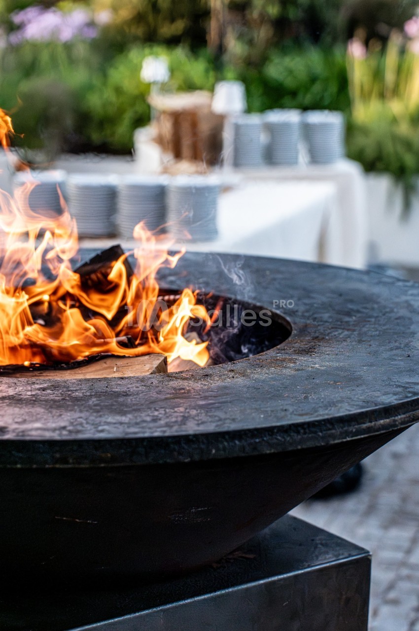 Brasero en métal avec des flammes vives, entouré d'une table dressée avec des assiettes empilées en arrière-plan.