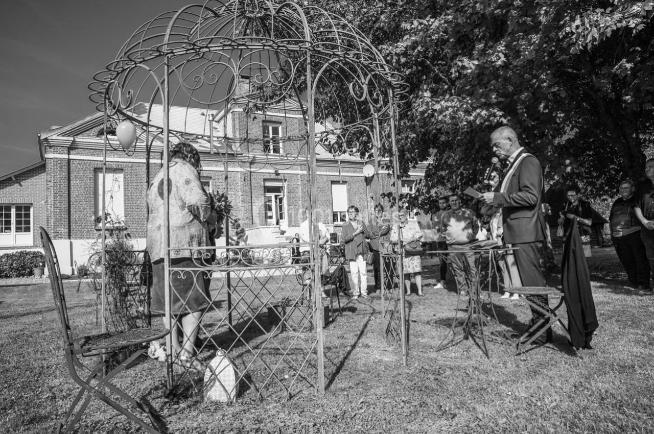 Cérémonie en plein air sous une structure métallique, avec des invités rassemblés devant un bâtiment ancien.