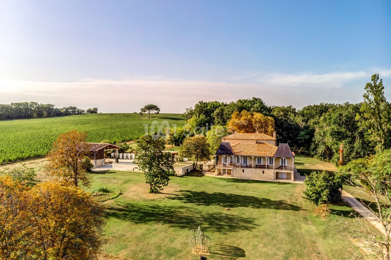 Vue aérienne d'une grande maison entourée de jardins, d'arbres et de champs sous un ciel dégagé.