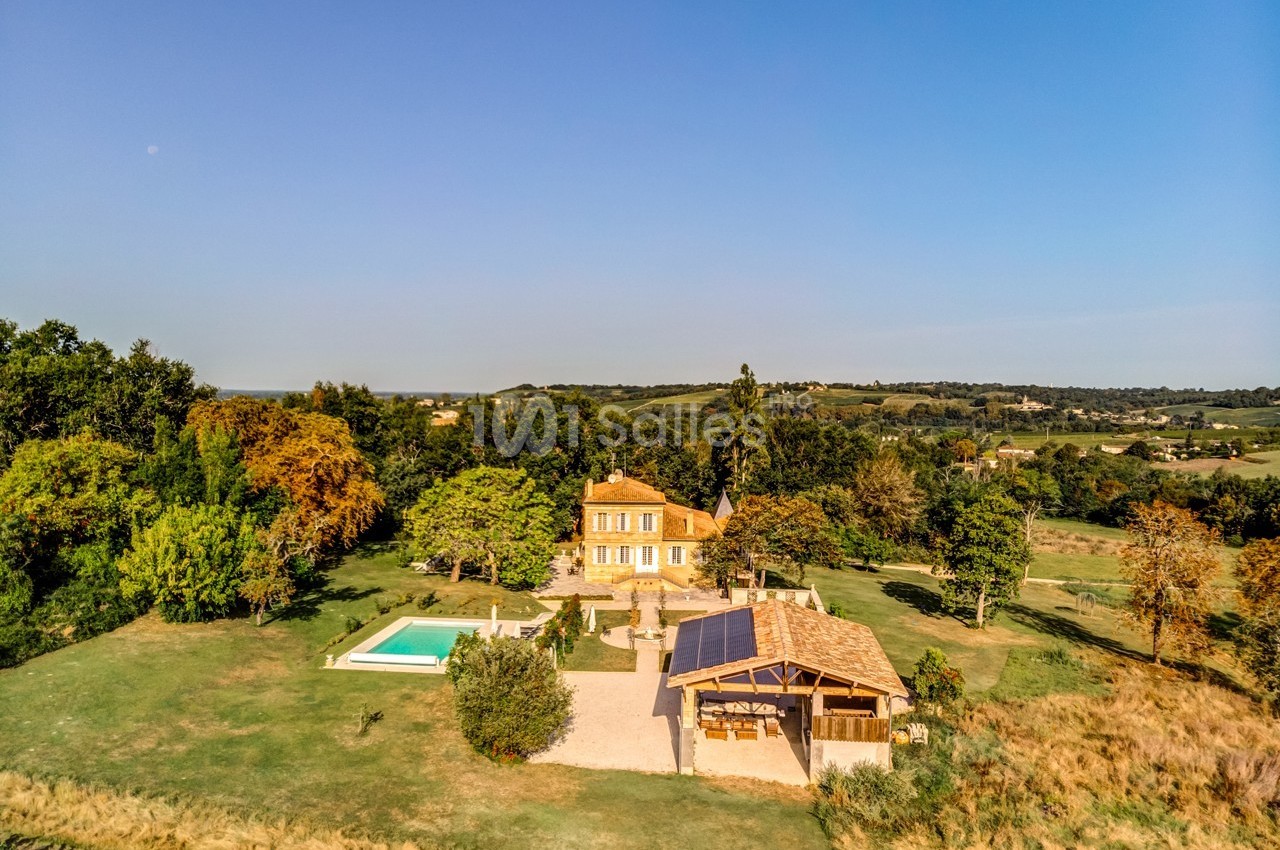 Vue aérienne d'une maison avec piscine, terrasse et jardin arboré, entourée de champs et collines sous un ciel dégagé.