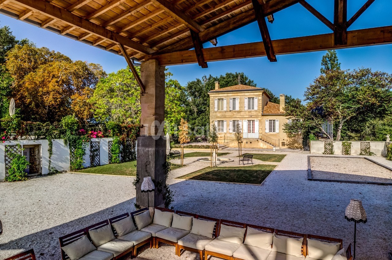 Vue d'une maison provençale entourée de jardins, prise depuis une terrasse couverte avec des sièges en bois.