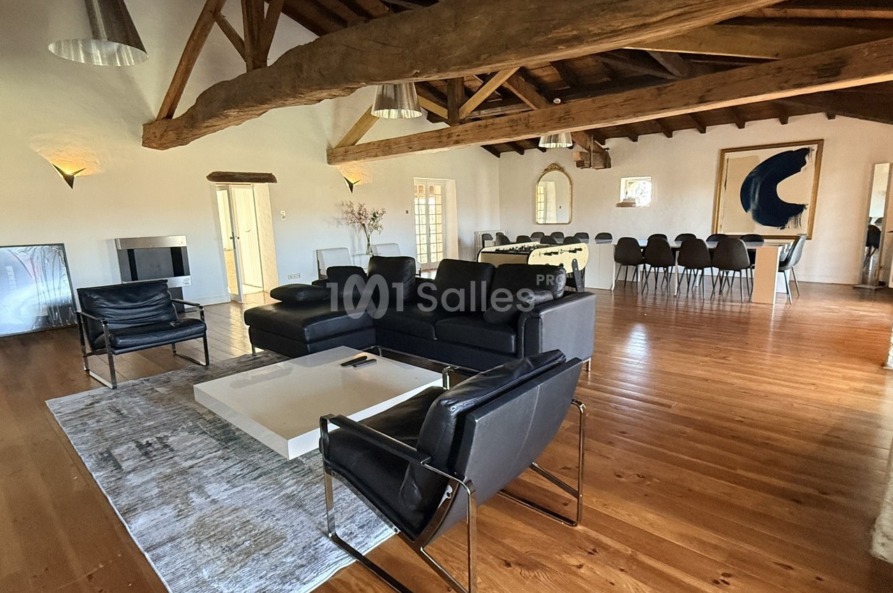 Salon spacieux avec poutres apparentes, canapé en cuir noir, table basse blanche et salle à manger en arrière-plan.