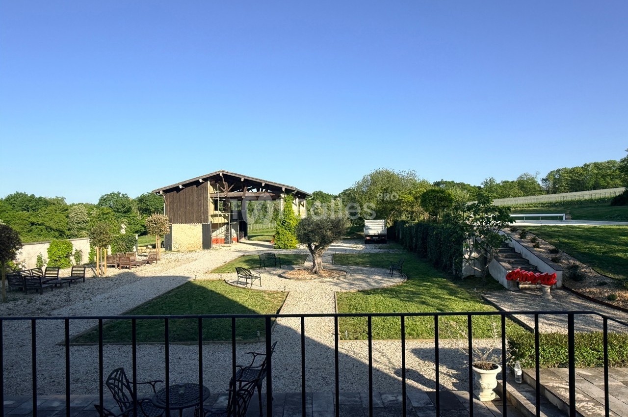 Vue d'une cour extérieure avec une grange, des espaces verts, des arbres et des meubles de jardin sous un ciel dégagé.