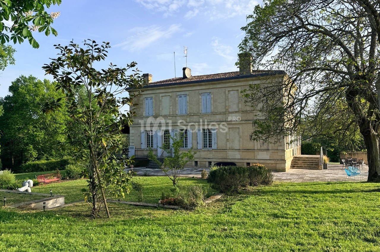Grande maison en pierre entourée d'un jardin verdoyant avec arbres, pelouse et mobilier d'extérieur.