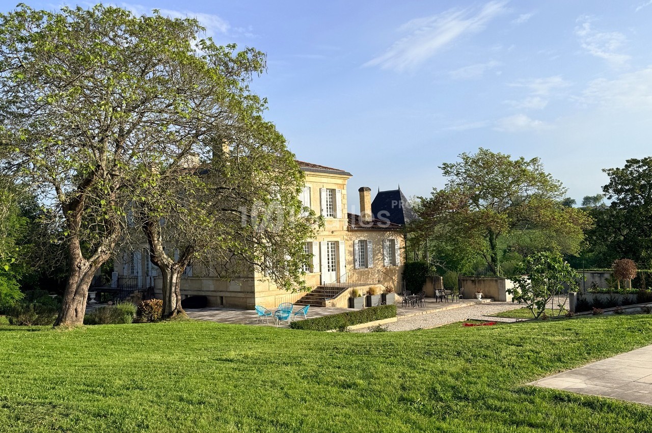 Manoir en pierre entouré de pelouse, arbres et terrasse, sous un ciel dégagé en journée.