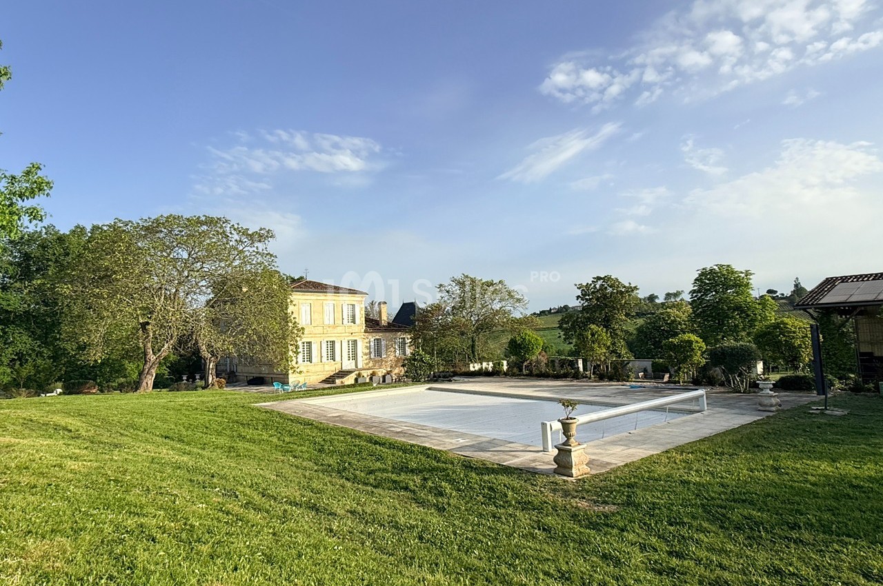 Grande maison en pierre entourée de verdure avec une piscine couverte au premier plan sous un ciel dégagé.