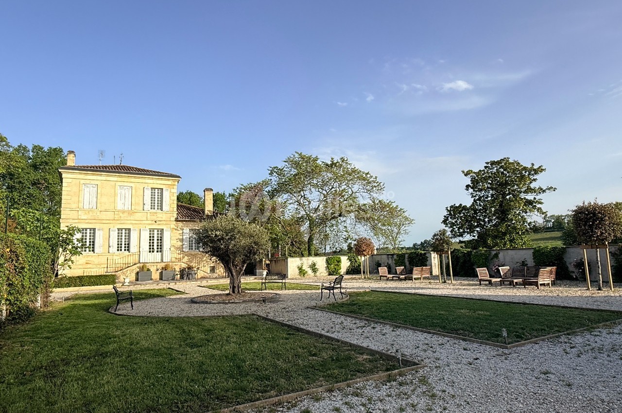 Jardin avec allées en gravier, pelouse, arbres et mobilier d'extérieur, devant une grande maison en pierre.