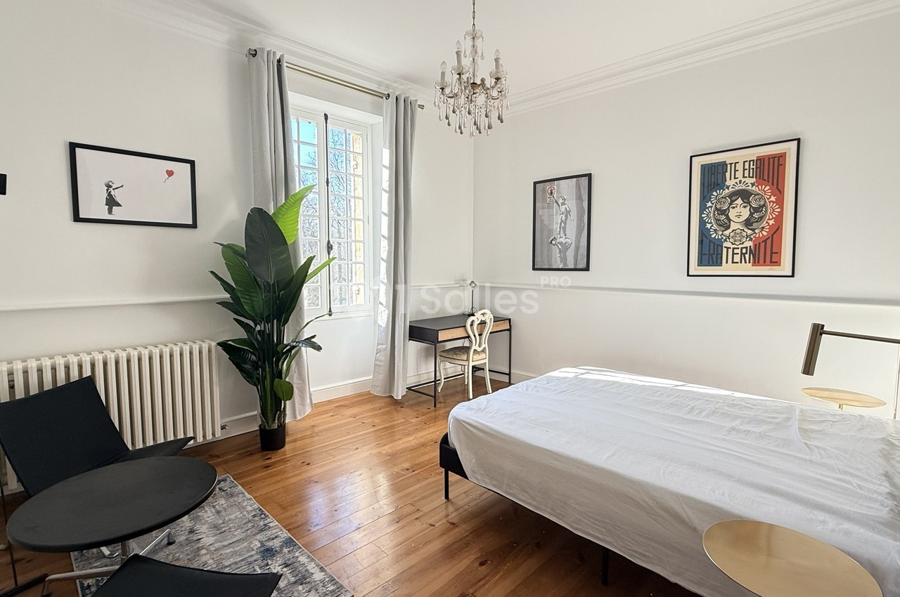 Chambre lumineuse avec lit, bureau, plantes, parquet en bois, tableaux encadrés et lustre suspendu.