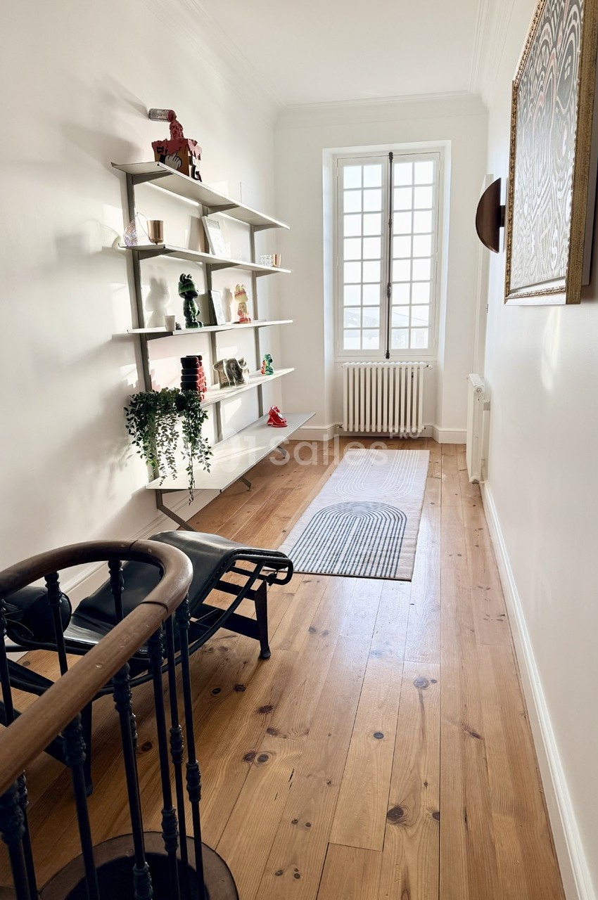 Couloir lumineux avec parquet en bois, étagères décoratives, chaise longue et fenêtre laissant entrer la lumière naturelle.