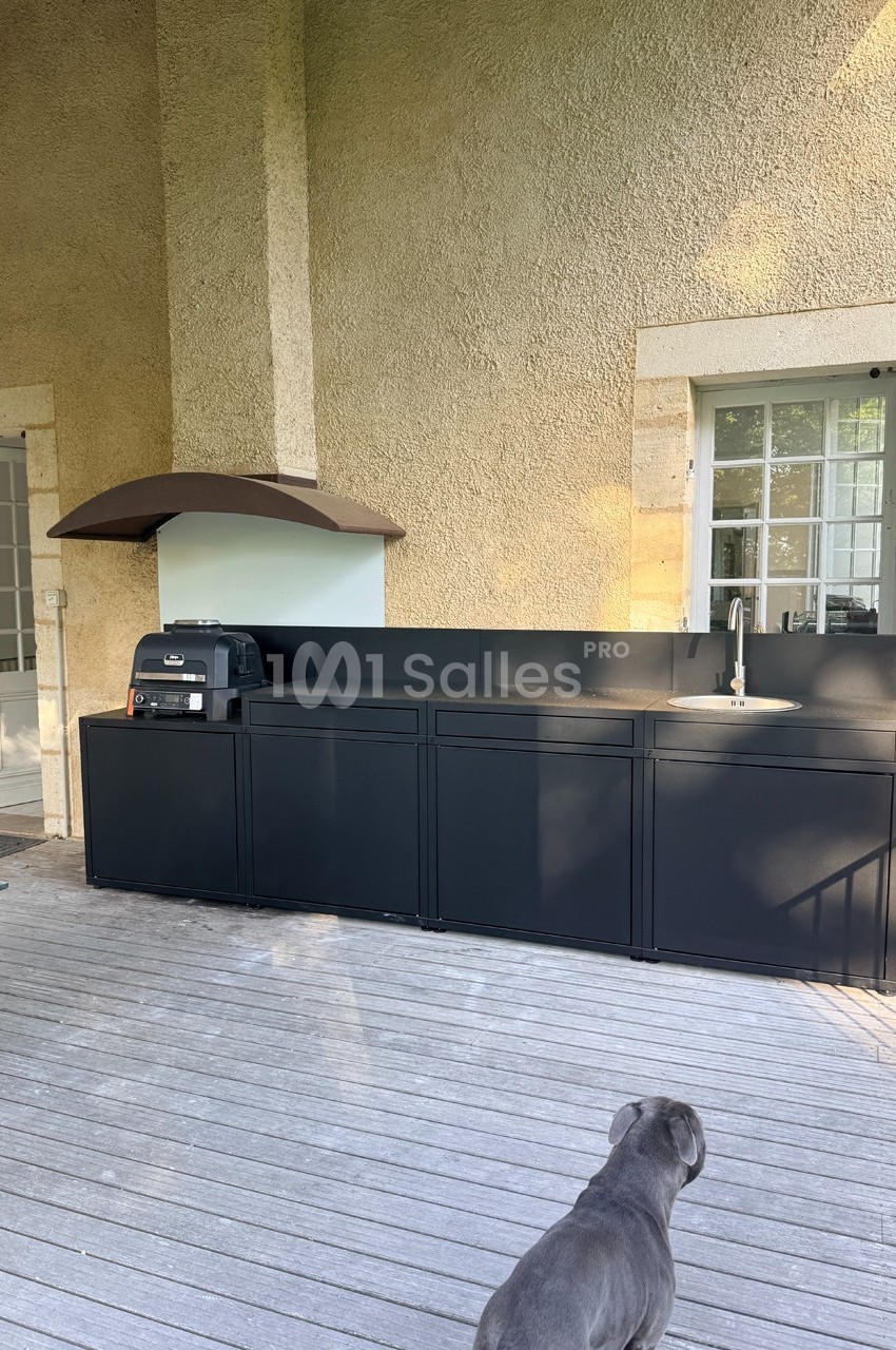 Cuisine extérieure moderne avec plan de travail noir, évier, barbecue et hotte, située sur une terrasse en bois.