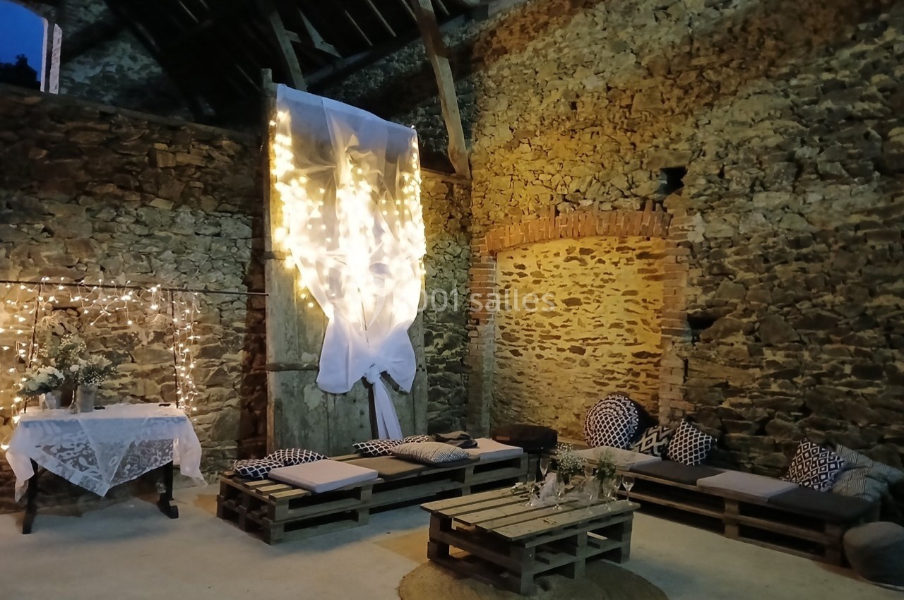 Espace de détente rustique avec murs en pierre, banquettes en palettes, guirlandes lumineuses et table décorée.
