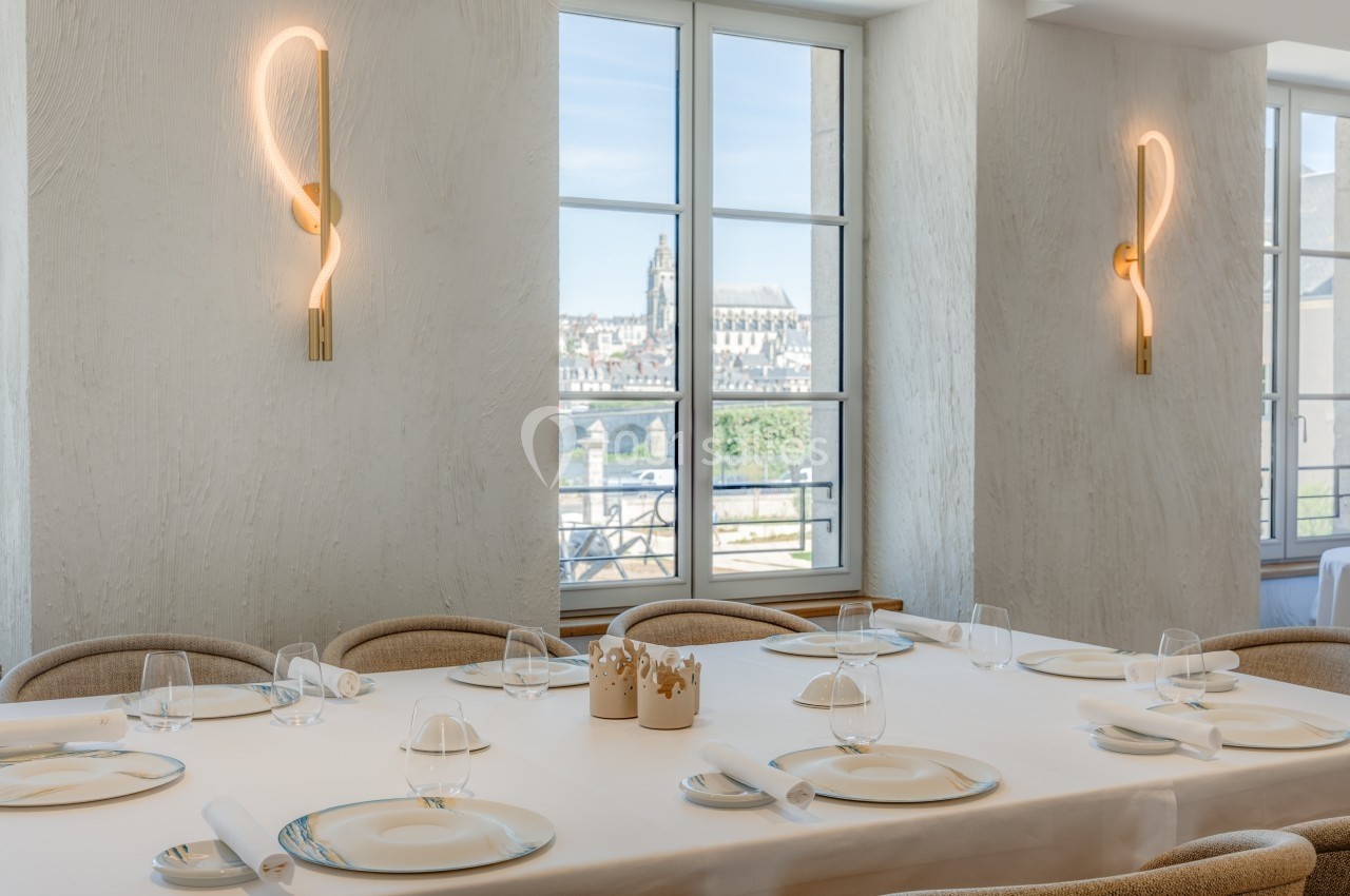 Salle de restaurant lumineuse avec tables dressées, grandes fenêtres et vue sur des bâtiments historiques à l'extérieur.