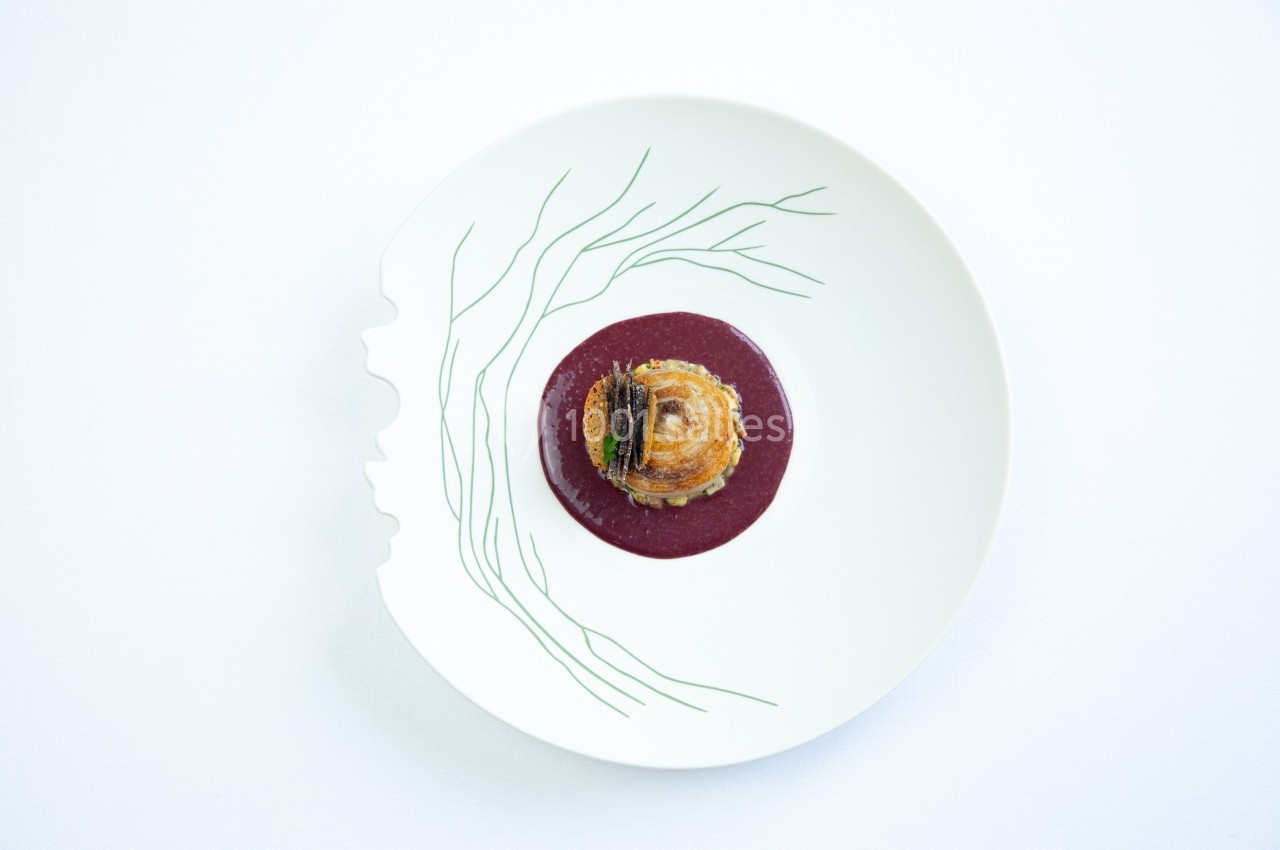 Assiette blanche avec un motif de visage stylisé, contenant un plat raffiné sur une sauce rouge.