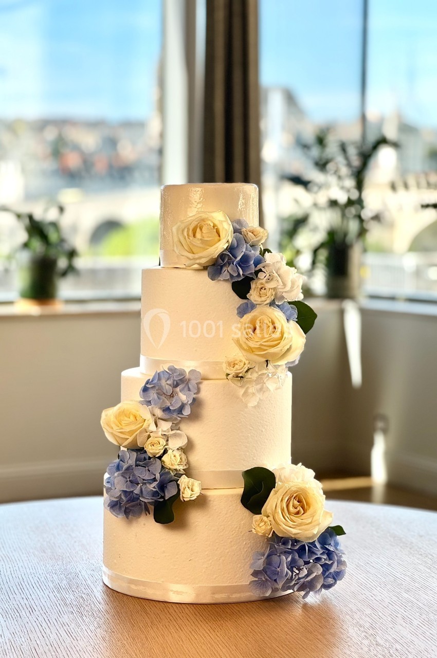Gâteau de mariage à trois étages décoré de fleurs blanches et bleues, posé sur une table devant une fenêtre.
