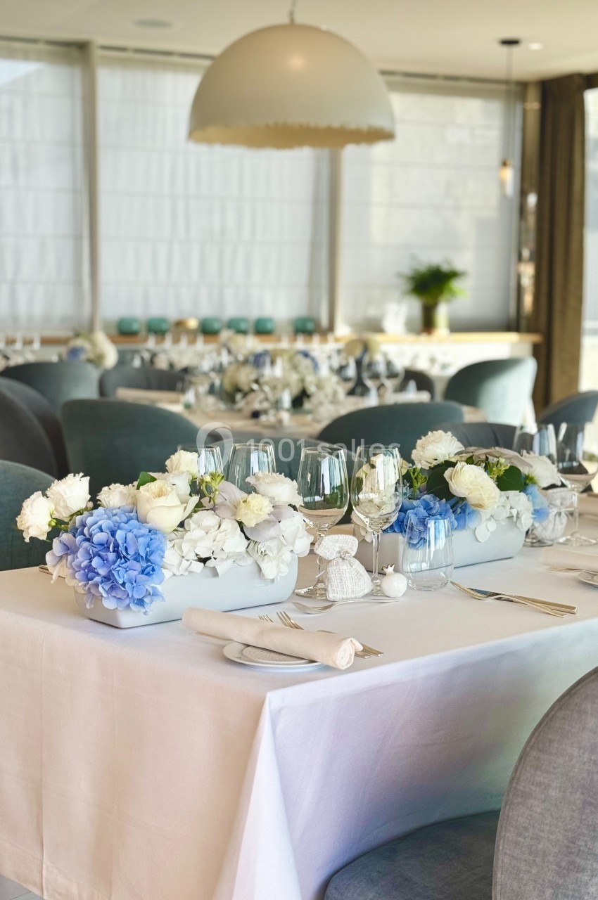 Salle de réception élégante avec tables dressées, décorées de fleurs blanches et bleues dans un cadre lumineux.