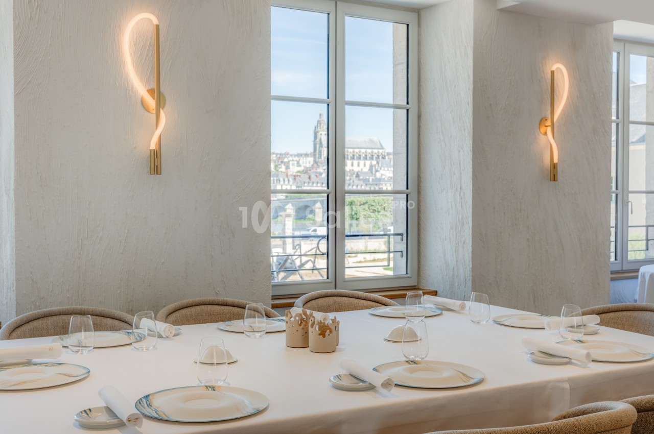 Salle lumineuse avec tables dressées, grandes fenêtres offrant une vue sur des bâtiments historiques et des appliques…
