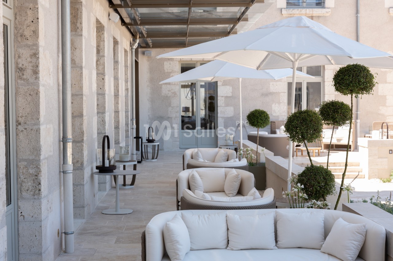 Terrasse élégante avec fauteuils blancs, parasols et plantes taillées, devant une façade en pierre claire.
