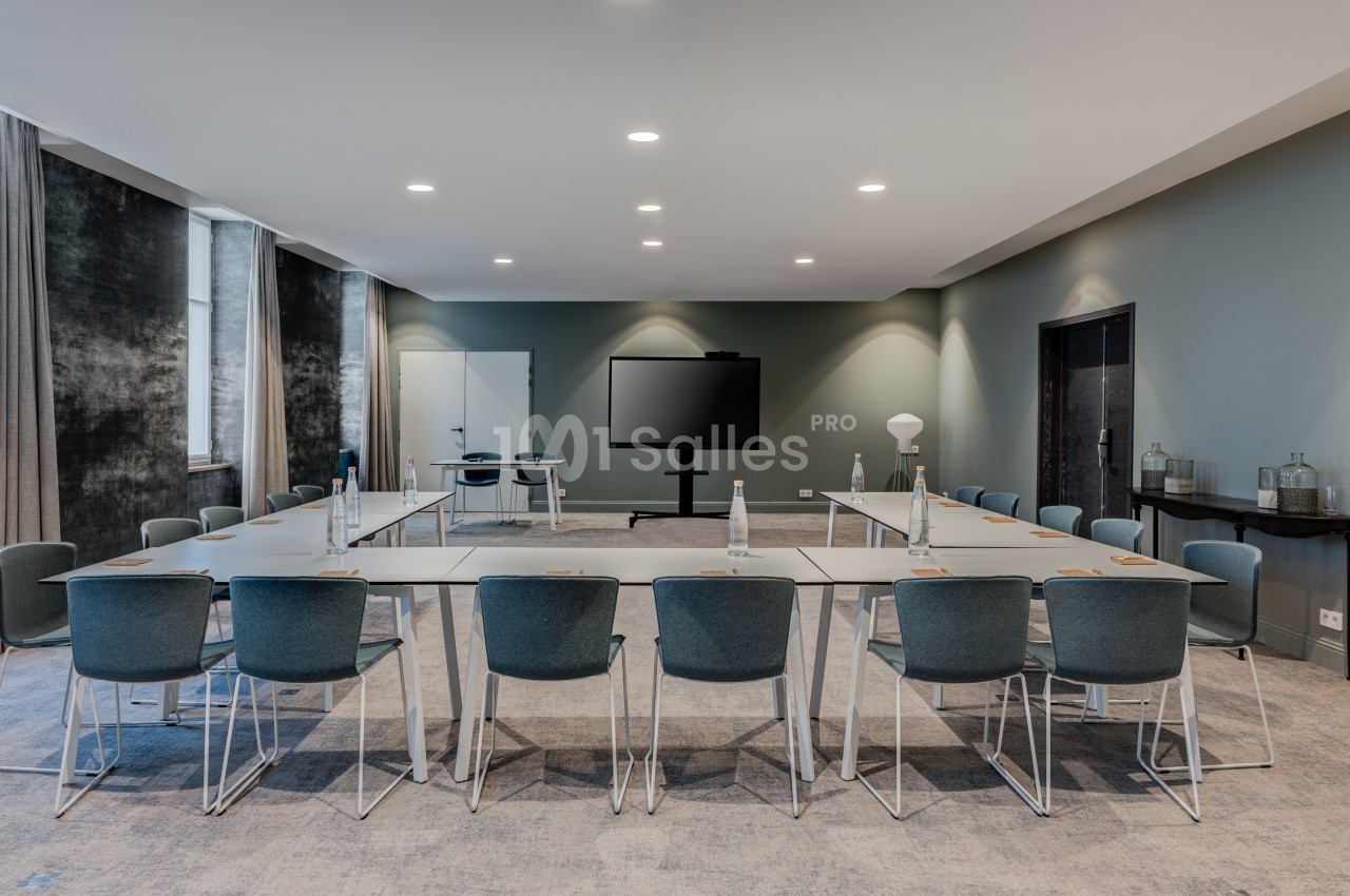 Salle de réunion moderne avec tables disposées en U, chaises bleues, écran de présentation et éclairage encastré.