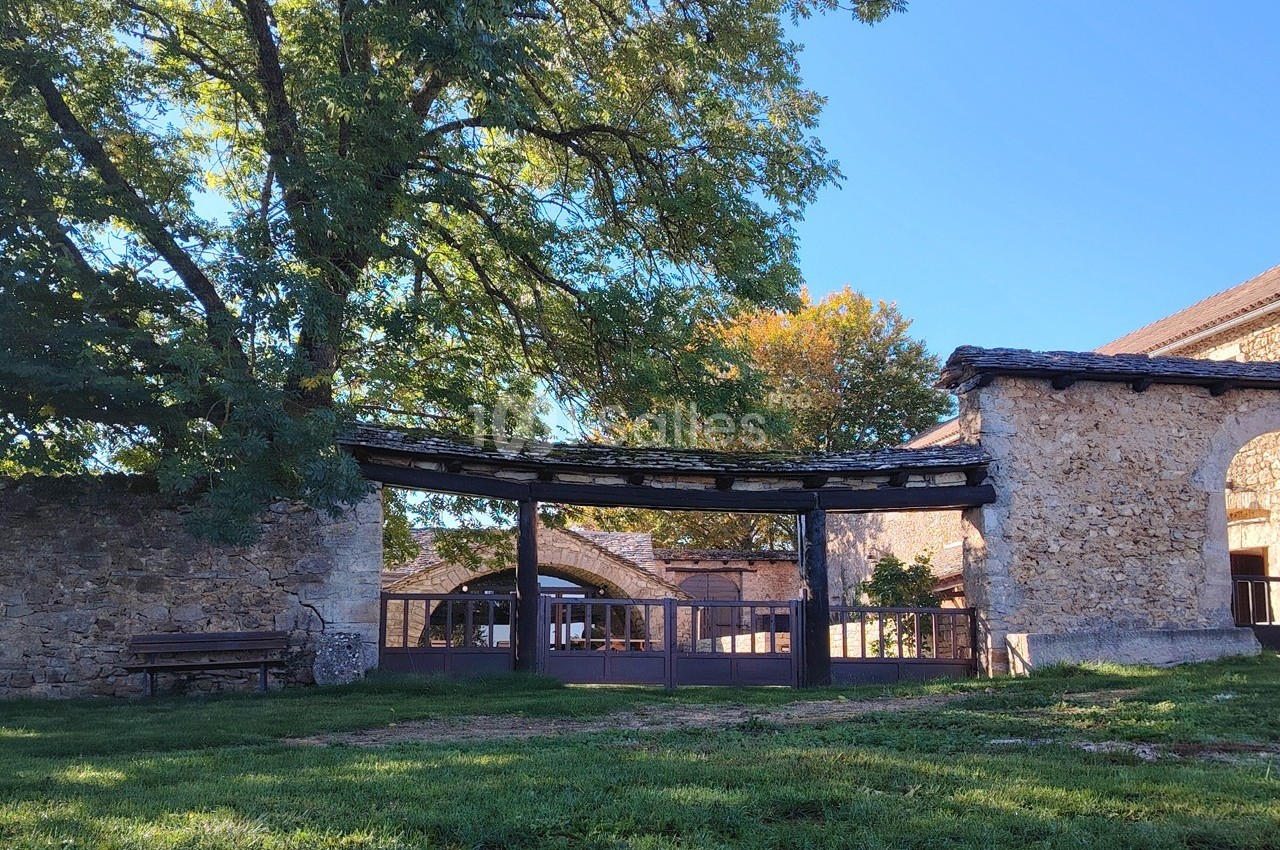 Entrée d'un bâtiment en pierre avec une arche en bois, entourée d'arbres et d'une pelouse verte.