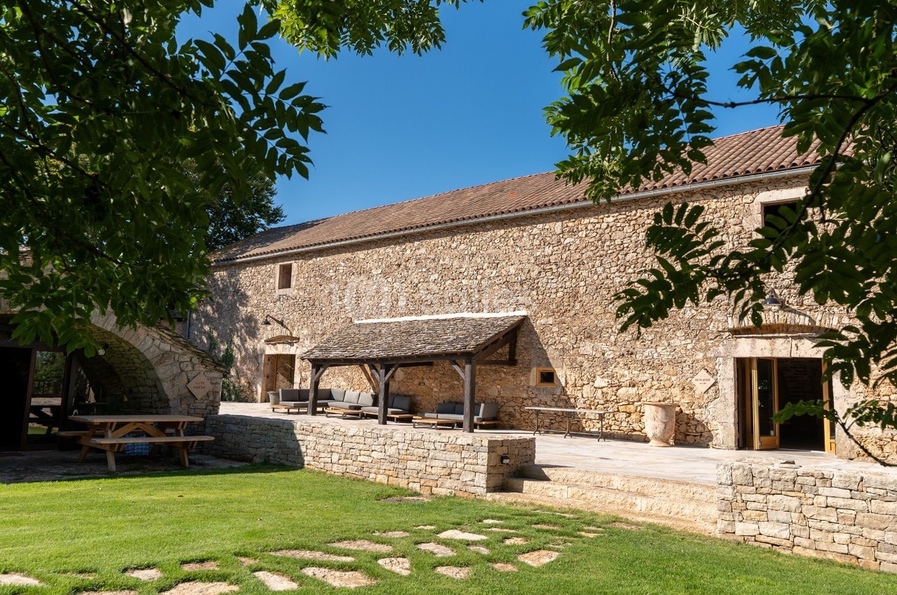 Bâtisse en pierre avec terrasse couverte, entourée de pelouse et d'arbres sous un ciel bleu.