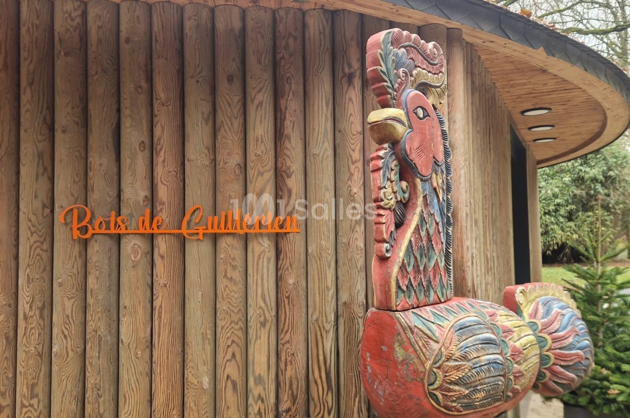 Sculpture colorée en forme d'oiseau devant un bâtiment en bois avec l'inscription ’Bois des Guillemins’.