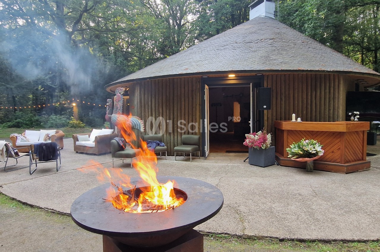 Brasero allumé devant une maison ronde en bois entourée de végétation, avec mobilier de jardin et décoration extérieure.