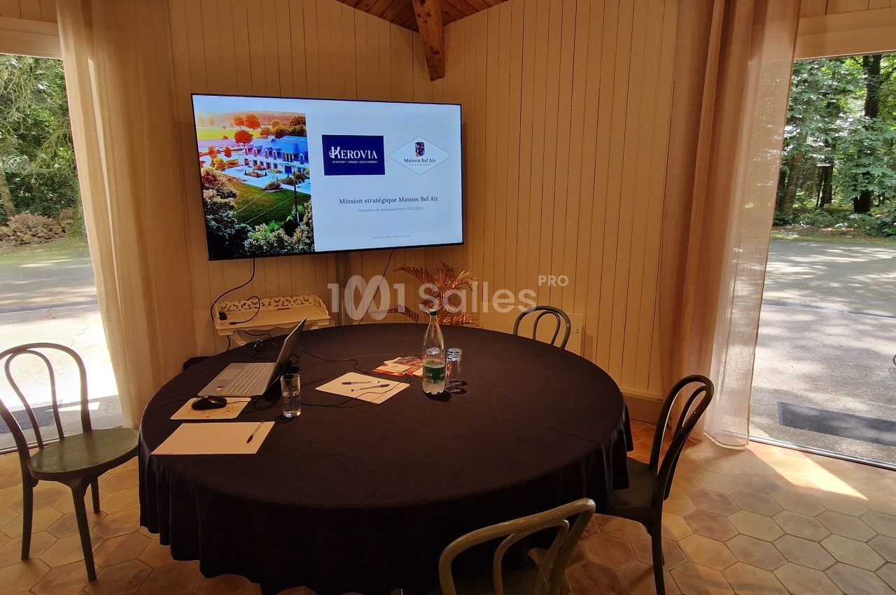 Salle lumineuse avec une table ronde, des chaises, un écran affichant une présentation et une vue sur un jardin boisé.