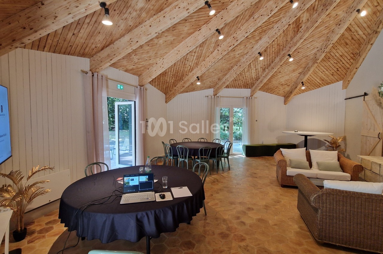 Salle lumineuse avec plafond en bois, tables rondes, canapé en osier, cheminée et écran de présentation.
