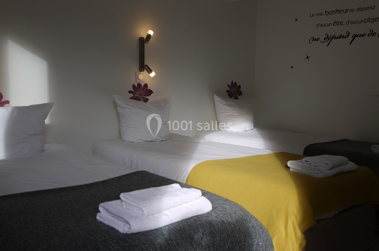 Trois lits simples avec couvertures colorées et serviettes pliées dans une chambre lumineuse décorée de citations murales.