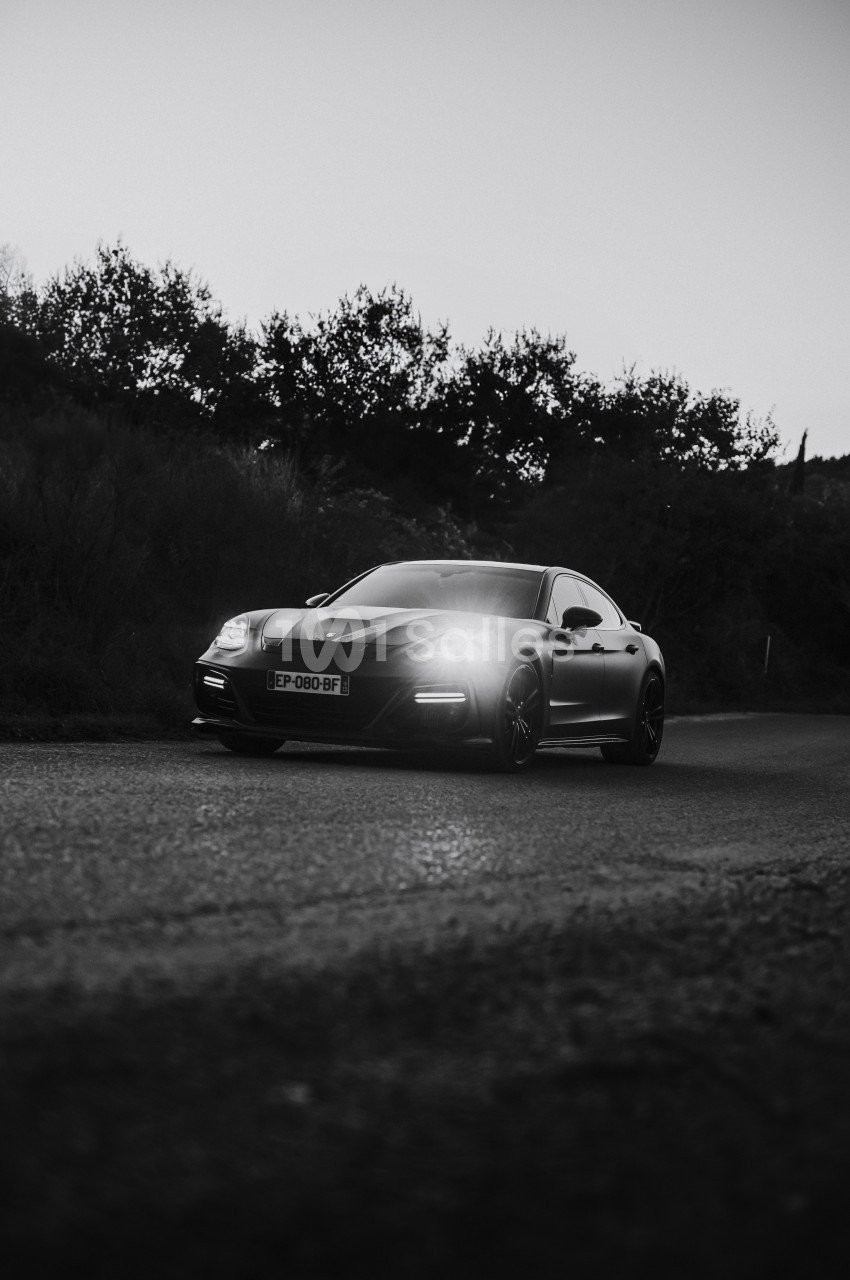 Voiture sportive noire stationnée sur une route, éclairée par ses phares, avec des arbres en arrière-plan.