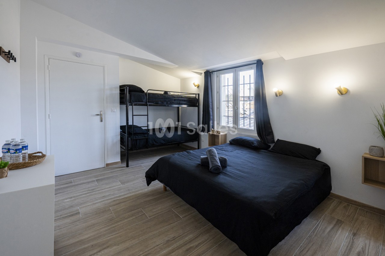 Chambre lumineuse avec un lit double, des lits superposés, parquet clair, murs blancs et une fenêtre avec rideaux.