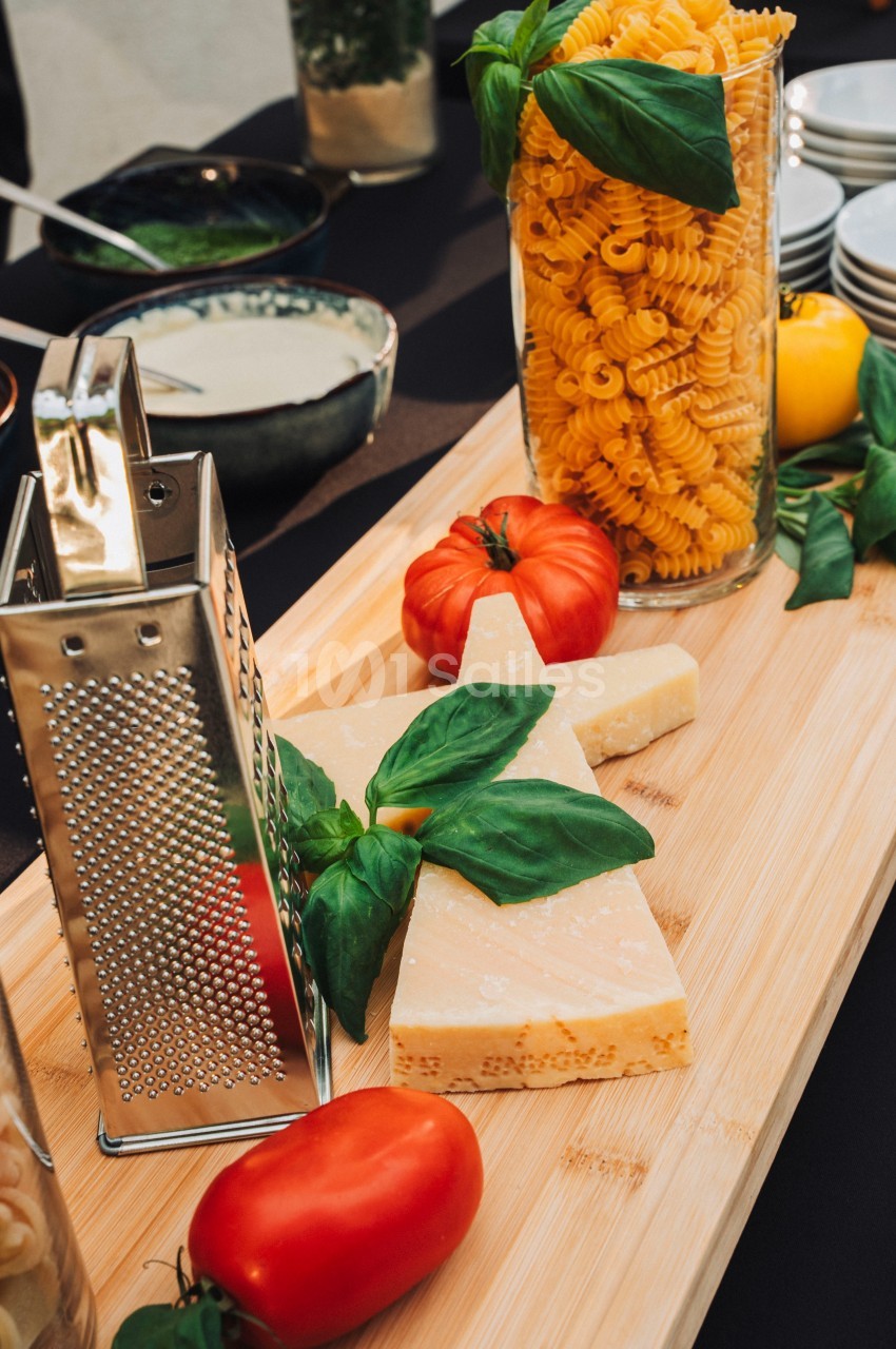 Planche en bois avec du parmesan, des tomates, du basilic, des pâtes en bocal et une râpe à fromage en métal.