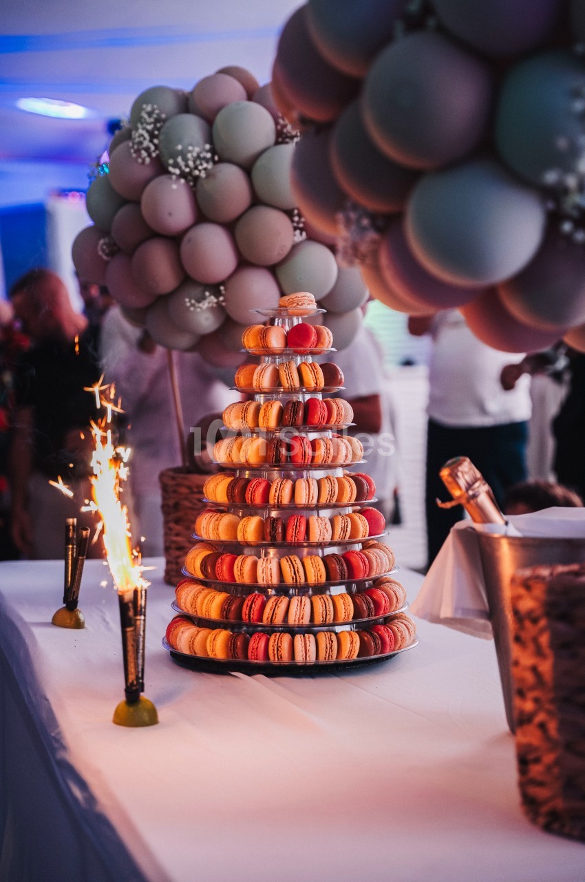 Tour de macarons colorés sur une table décorée, entourée de ballons et de fontaines étincelantes.