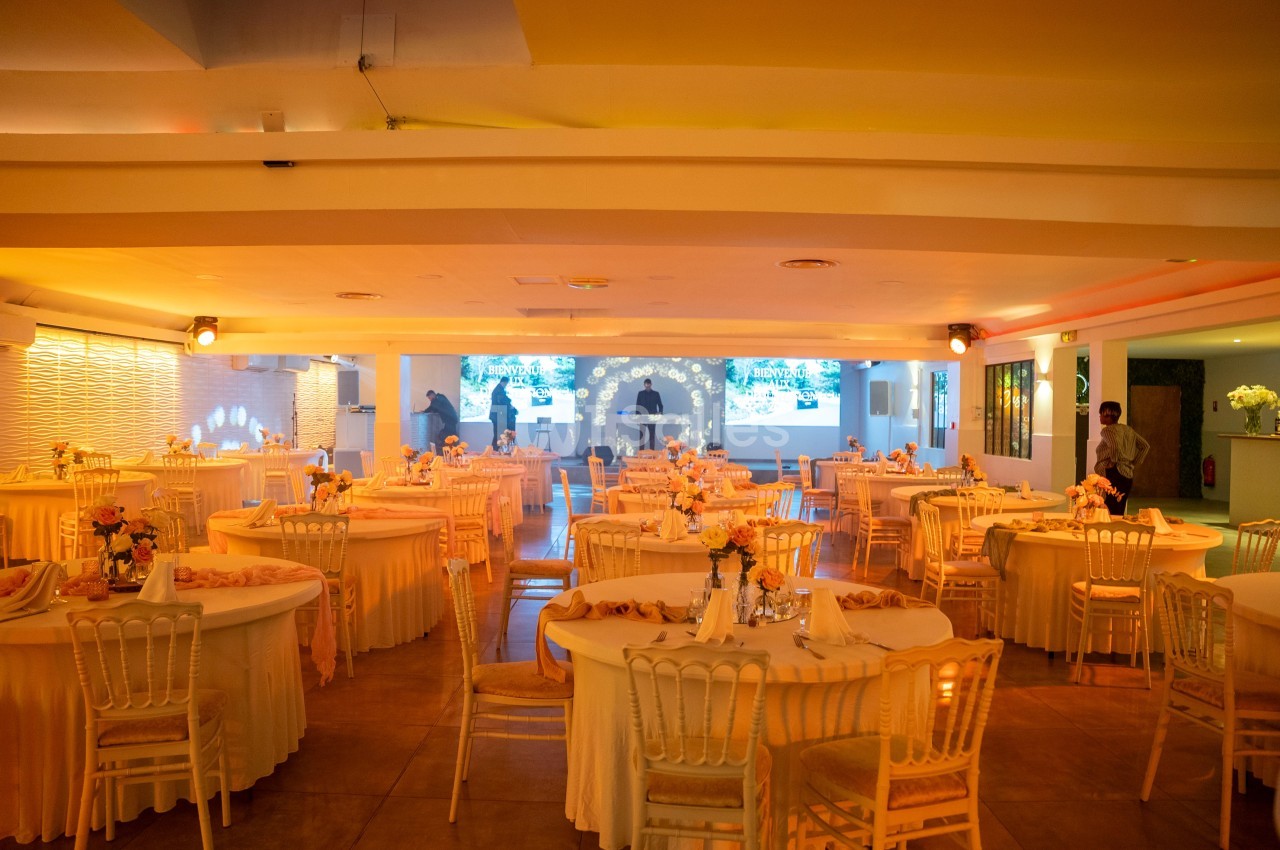 Salle de réception lumineuse avec tables rondes décorées de nappes et fleurs, chaises blanches et éclairage chaleureux.