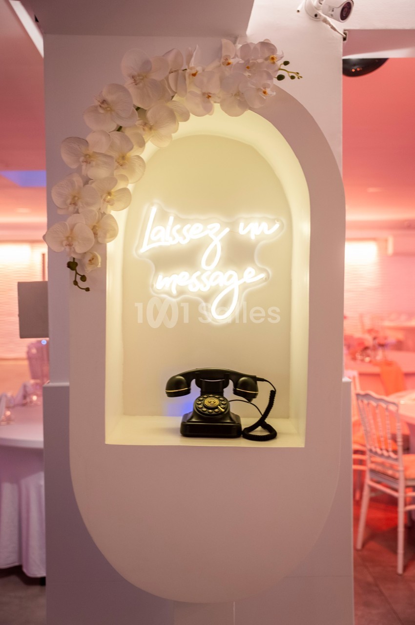 Téléphone vintage noir sous une enseigne lumineuse ’Laissez un message’, encadré par des orchidées blanches.