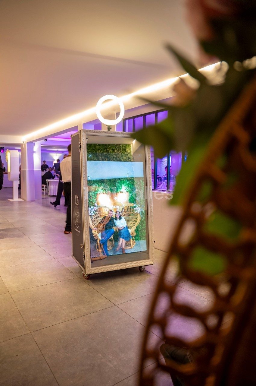 Miroir interactif dans un cadre lumineux, reflétant une scène avec des fauteuils en osier et un décor végétal.