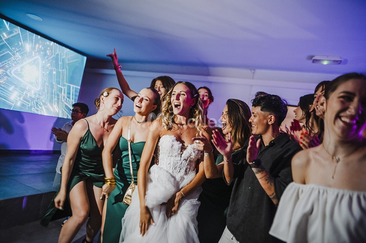 Un groupe de personnes, dont une mariée en robe blanche, dansant et célébrant dans une ambiance festive en intérieur.