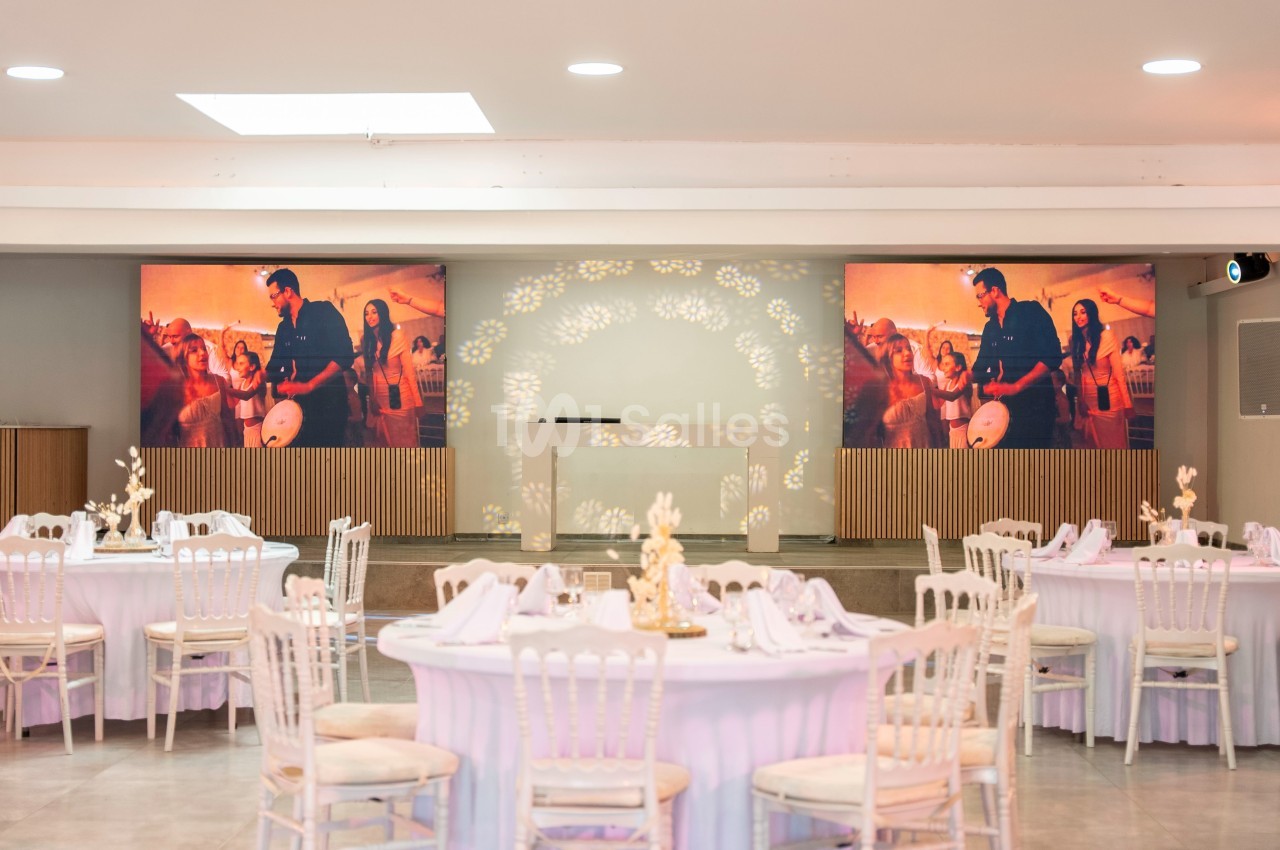 Salle de réception décorée avec des tables rondes élégantes et écrans affichant une scène festive en arrière-plan.