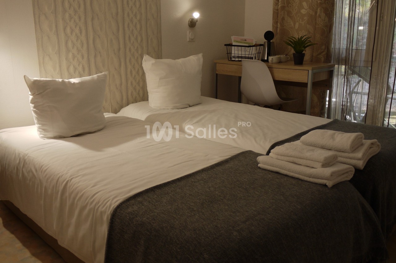 Deux lits simples avec draps blancs, couvertures grises, serviettes pliées, bureau et chaise près d'une fenêtre.