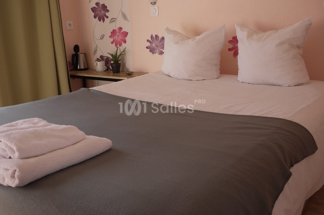 Lit double avec draps blancs, couverture grise, serviettes pliées et décoration murale florale dans une chambre lumineuse.