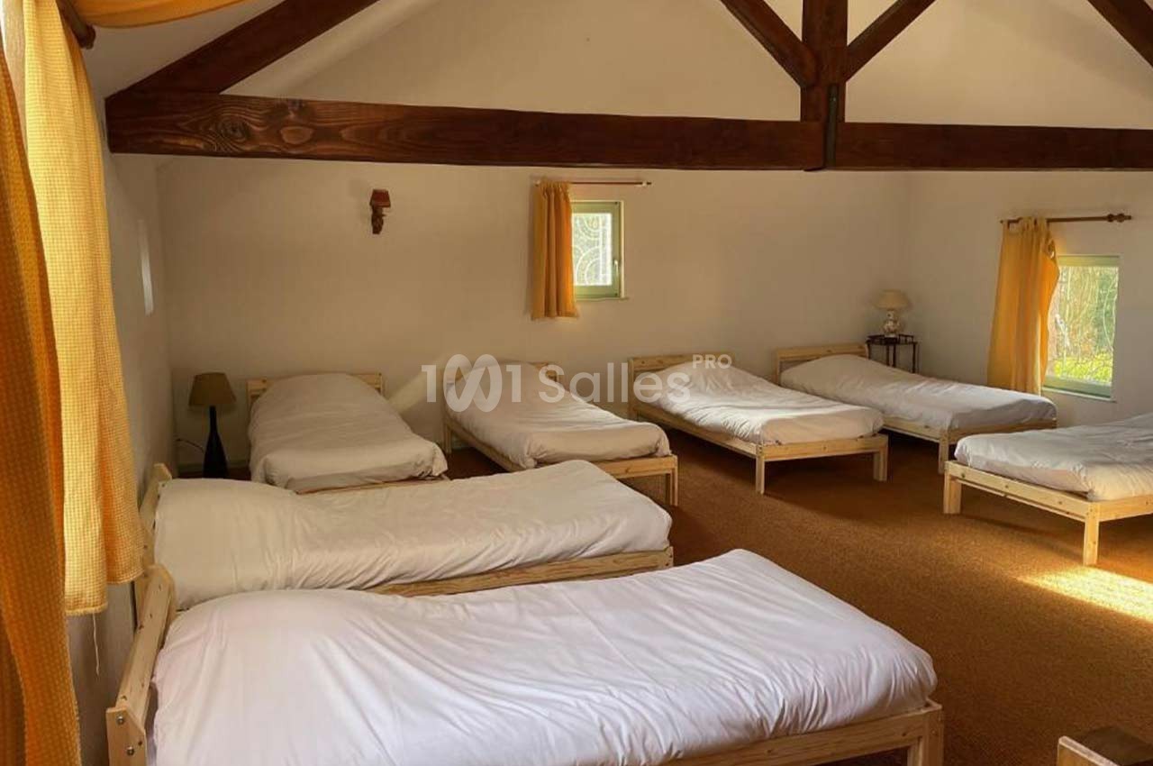 Dortoir lumineux avec six lits simples en bois, draps blancs, poutres apparentes et fenêtres avec rideaux jaunes.